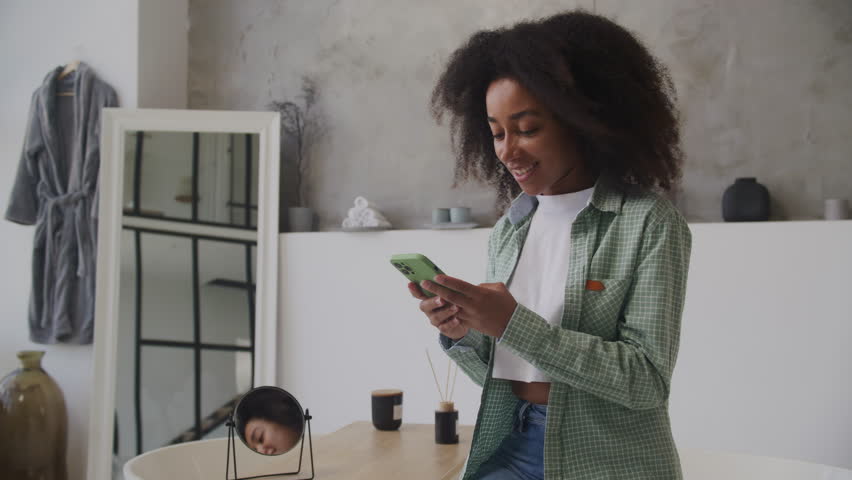 African American Woman Browsing her Smartphone at Home. Black female texting using cell phone sitting in bathroom. | Shutterstock HD Video #1109082159