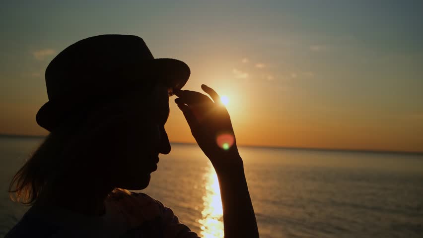 Silhouette of inspired woman in a hat at sunset by the sea. Female profile on the background of setting sun. Live camera movement.