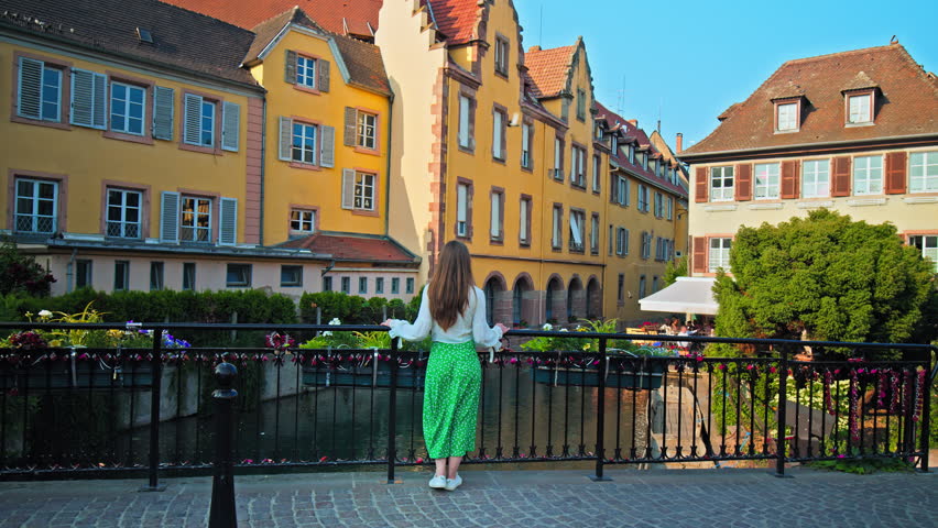 Beautiful Girl on a bridge looking at colourful Timber framing houses in Colmar. A female tourist in an elegant dress enjoying the view of Little Venice in Colmar Town, Grand Est, France Royalty-Free Stock Footage #1109082381