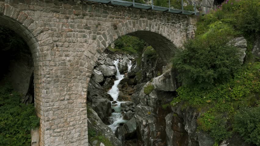Flying under arch of brick bridge while river water stream is flowing in summer season in Furka Pass valley of Switzerland. Aerial drone view
