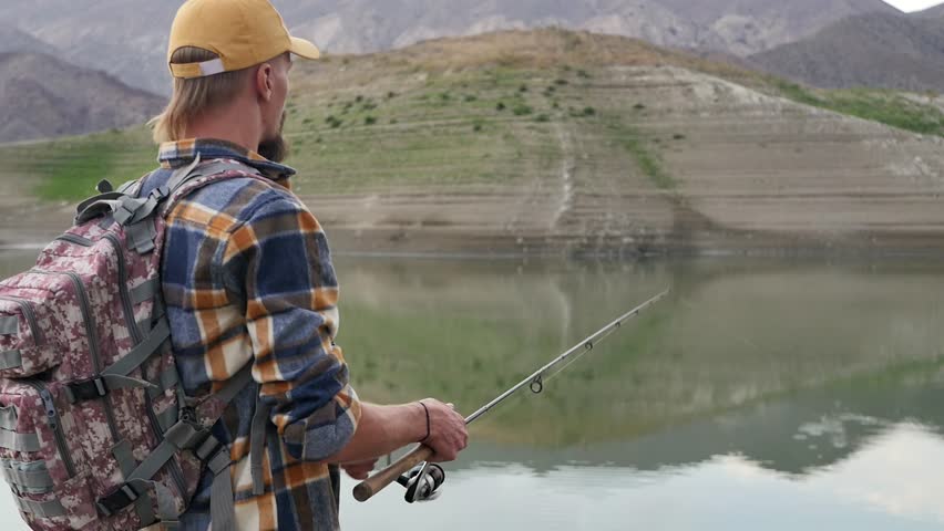 A Caucasian young handsome fisherman reels in a fishing rod while standing on the shore of a mountain lake and tries to catch a fish using a spinning rod to catch predatory fish. Fishing alone