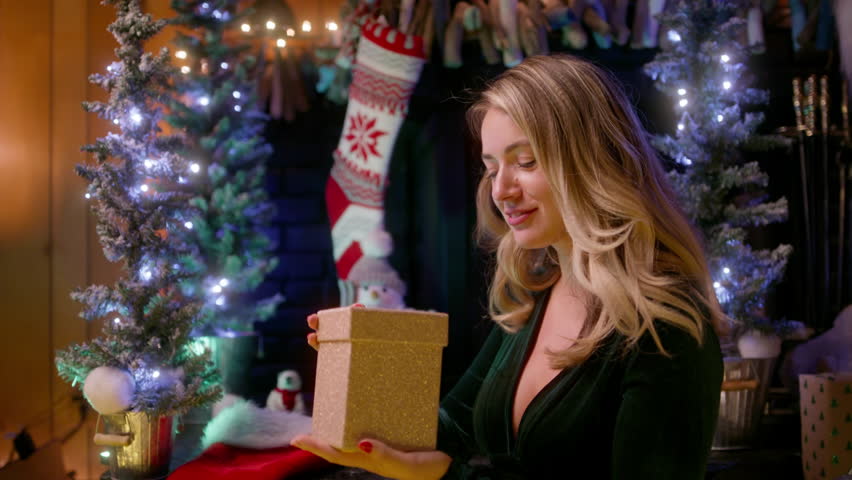 Gorgeous young beautiful woman opening a golden gift box, light shines on her beautiful face. Attractive smiling model wears a velvet emerald dress, looks happy. Christmas tree lights on background 4K Royalty-Free Stock Footage #1109093495