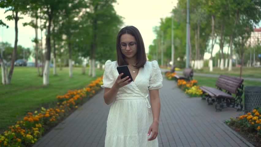 A young woman walks along a flower alley with a phone in her hands. A girl in a white dress looks at her phone and walks among the flowers. | Shutterstock HD Video #1109094843