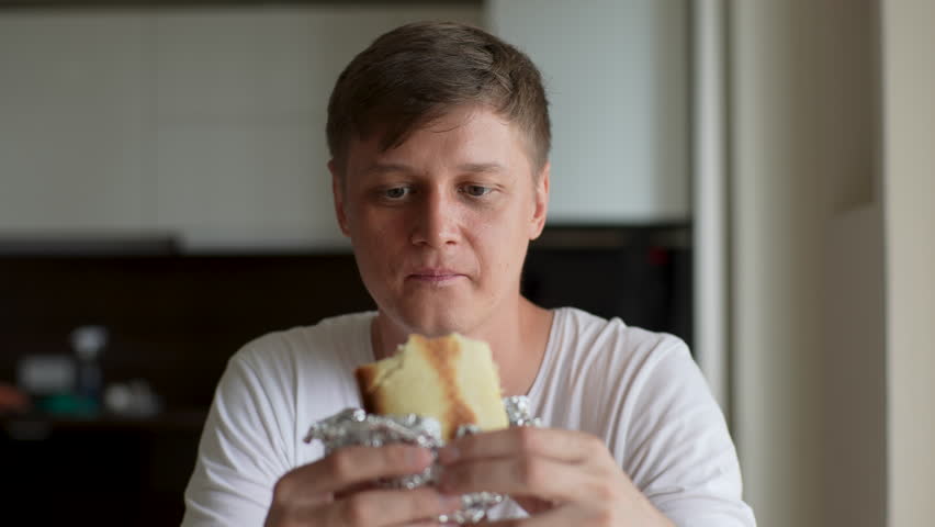 Close-up portrait of disgruntled young man eating unappetizing sandwich at kitchen room. Hungry handsome male disappointed with taste of bad quality fast food at home. Shooting in slow motion.