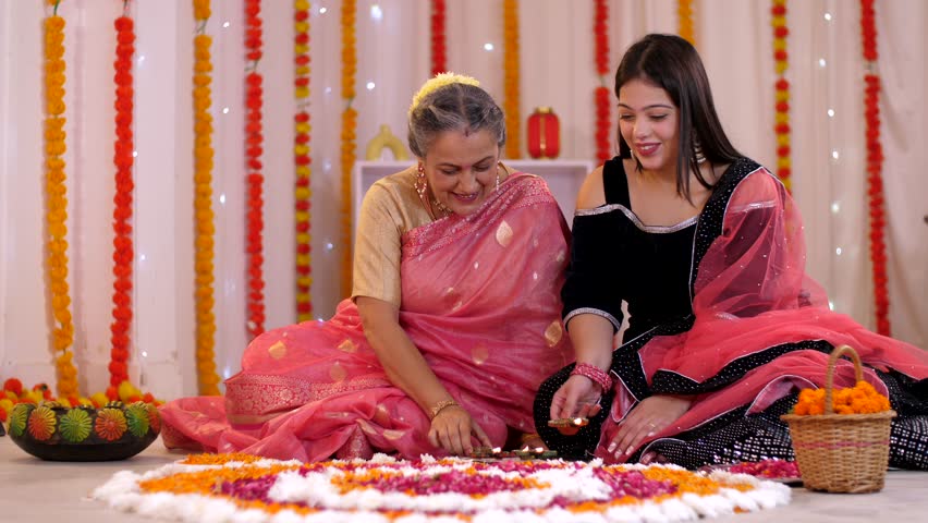 Pan shot of a mother and daughter decorating a traditional Rangoli with Diyas. Pretty young girl and her mother sitting on the floor while decorating the house for Diwali celebrations - a Hindu fes...
