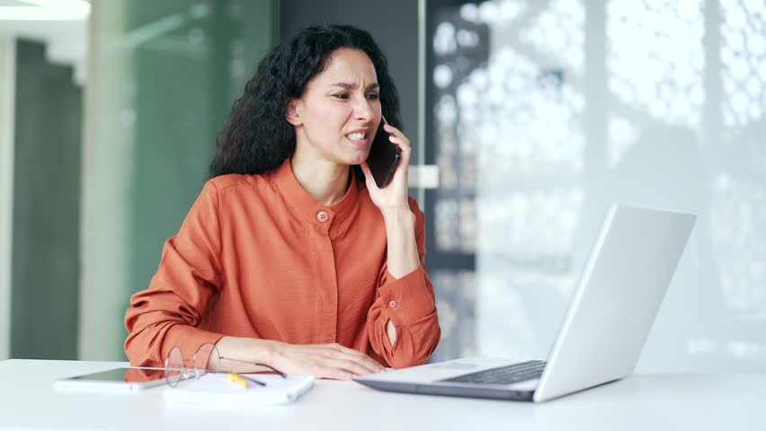 Angry female employee arguing talking on mobile phone while sitting at desk at workplace in modern office. Frustrated curly brunette woman argues, regrets explaining the problem using a smartphone | Shutterstock HD Video #1109109363