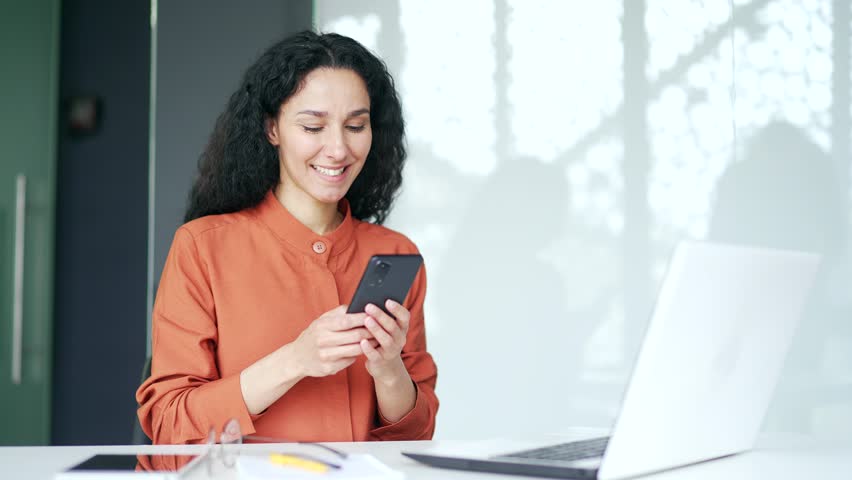 Young female employee is using smartphone while sitting at workplace in modern office. Smiling curly brunette woman typing message, chatting online, scrolling, browsing social media or checking e-mail | Shutterstock HD Video #1109109395