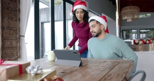 Happy biracial couple wearing santa claus hats using tablet for video call at home, in slow motion. Lifestyle, celebration, christmas, communication and domestic life, unaltered.