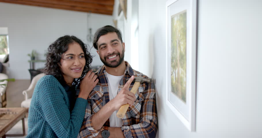 Happy biracial couple looking at painting on wall at home, in slow motion. Lifestyle, renovation and domestic life, unaltered. Royalty-Free Stock Footage #1109114103