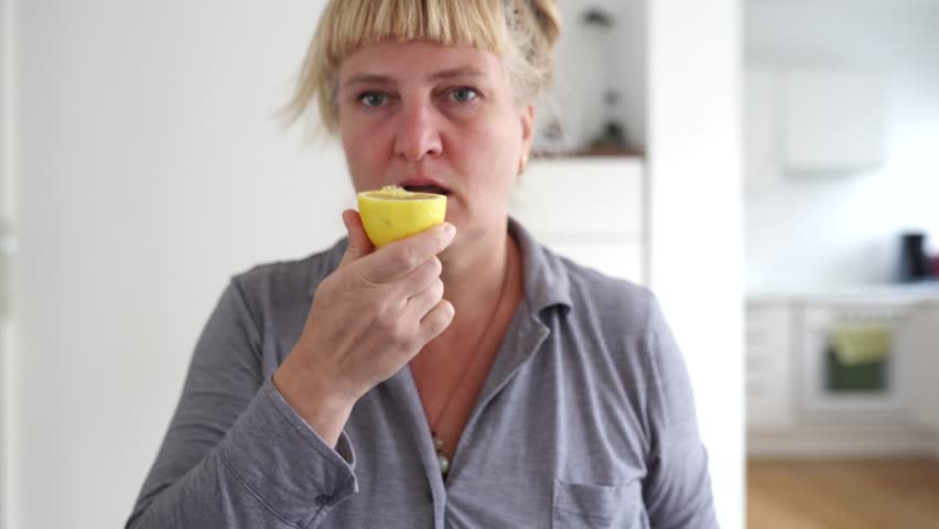 middle-aged woman, sitting table in home dining room, eats sour ripe lemon, grimaces, shudders from taste, lemon sour face. woman bites ripe lemon and makes face