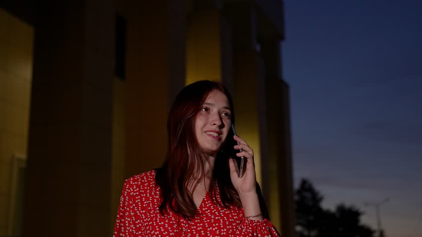 Happy young woman talks on phone walking along night city street. Smiling woman has phone conversation while walks across evening town. Young woman talks with boyfriend on phone on dark street | Shutterstock HD Video #1109116161