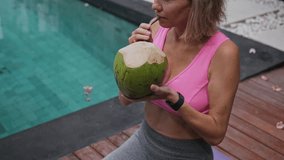 Mid adult woman has a rest after yoga class and drinking green fresh coconut near the swimming pool