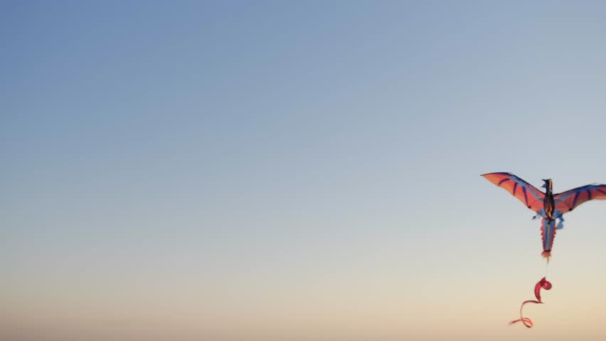 A kite in the form of a red bird soars high in the sky, smoothly gliding in the air against the background of the blue sky in the sunset rays.