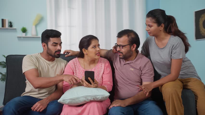 Indian Middle aged mother ordering food for family on mobile phone while by asking adult sibling kids and husband on sofa - concept of cyberspace, home lifestyle and Technology.