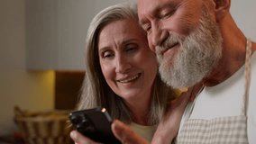 Close up happy old senior Caucasian family laughing in kitchen bonding mature couple grandparents using funny mobile app read news smiling retired man husband and woman wife shopping online on phone