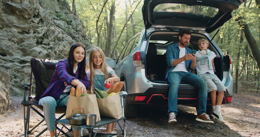 Happy family having weekend picnic outdoors in the forest. Woman and girl preparing food and father and son siting in open trunk of car and using phone. Time to Travel concept. Family travel dream Royalty-Free Stock Footage #1109135927