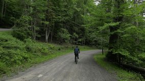 man riding bicycle down hill filmed from behind (footage from back, casual clothes, mountain bike, gravel path) descent, aerial footage, following descending rural road with trees