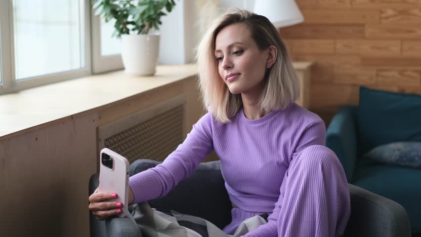 Happy young woman 20 years old in casual clothes, purple home suit sitting on a sofa indoors, relaxing at home in her own apartment room. The blonde girl is holding a smartphone in her hands and | Shutterstock HD Video #1109137913