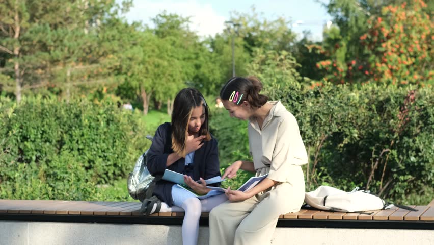 Cute happy little schoolchildren with backpacks and notebooks sitting on a park bench outdoors. Two young schoolgirls doing homework outdoors