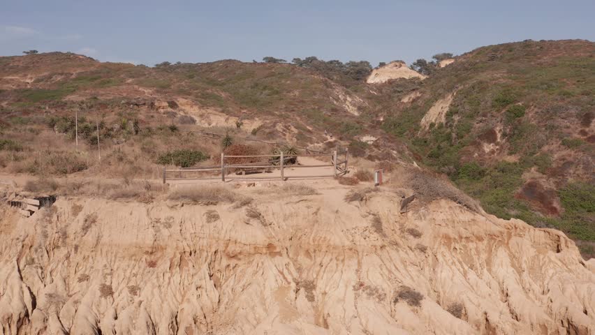 Footage circling La Jolla Shores cliff lookout. The top of the cliff has grass and shrubs. The cliff is steep and sandy brown. The ocean is deep and blue and it is day in the afternoon.