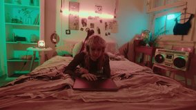 Young girl in wireless headphones opening laptop, getting online message, smiling and typing an answer while lying on bed in 90s aesthetic room with neon light. Whip zoom shot - Powered by Shutterstock - Get 15% off with code: PIKWIZARD15