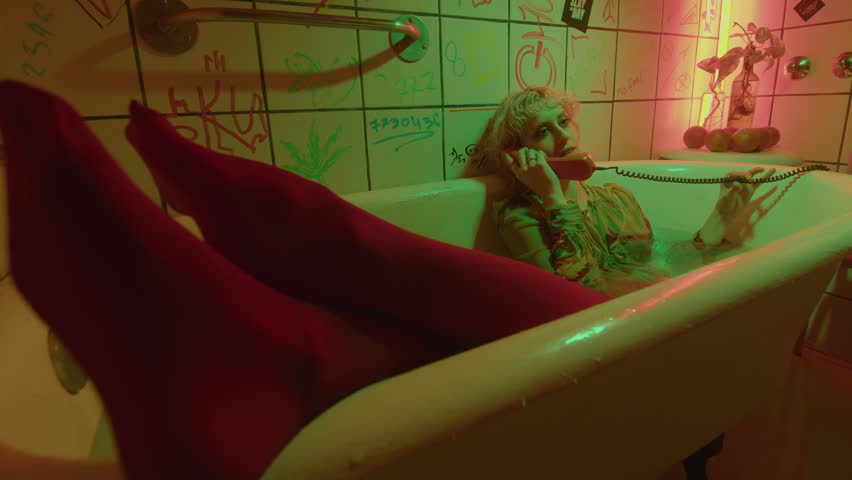 Young relaxed girl in red tights lying in bathwater with feet close to the camera and speaking on a landline phone in bathroom with neon illumination and drawings on tile