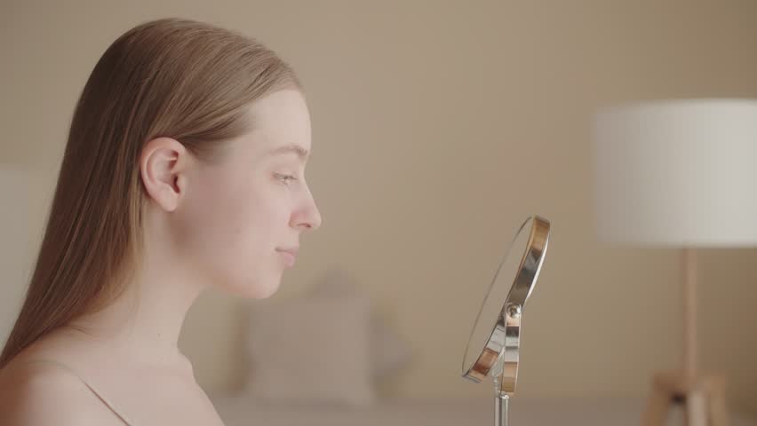 Satisfied attractive young lady looks in reflection mirror, enjoying effect after the applying of the cosmetic product. Caucasian young woman with perfect skin. Skin care advertisement. Slow motion Royalty-Free Stock Footage #1109146277