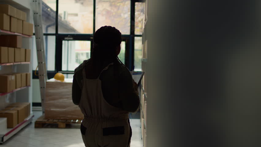 Dolly in tracking shot of logistics director inputing updated products info online, walking through facility aisles surveying labels on wares while assistant scans boxes in background