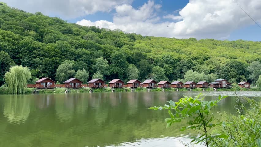 Beautiful landscape of small wooden holiday houses for tourists near a lake and green forest on a hillside in summer on a sunny day. Recreation complexes in the mountains near the lake.