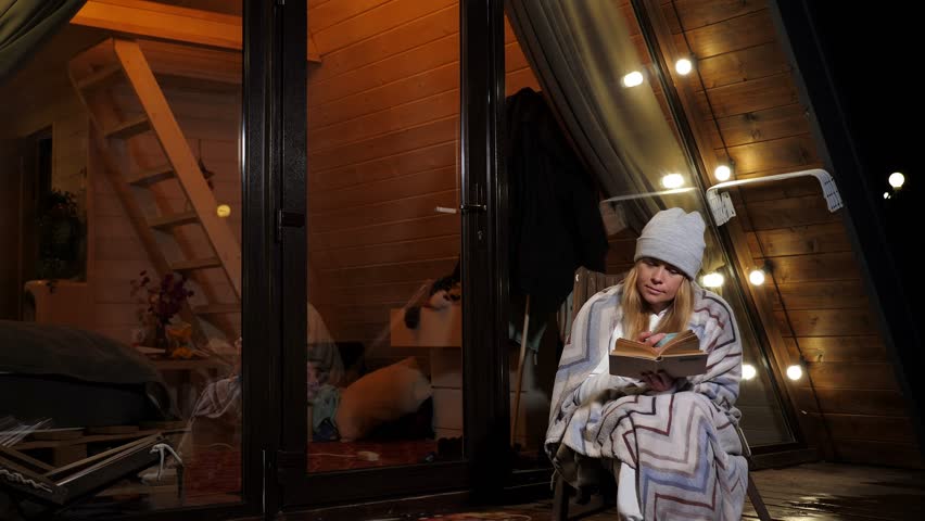 A woman in a hat with a plaid is sitting in an armchair with a cup of tea and a book in the evening, relaxing in a country triangular house.The house is a bungalow with a roof descending to the ground