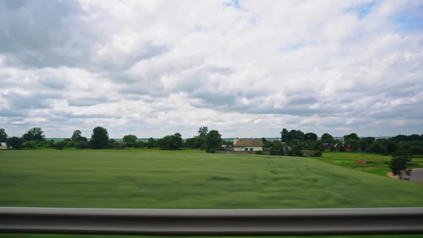 Road along village with cottages in rural area. Traffic barrier guard rail along highway. View through side window of van in motion. Roadway out of town. Meadows fields. Cloudy weather, overcast Royalty-Free Stock Footage #1109168549