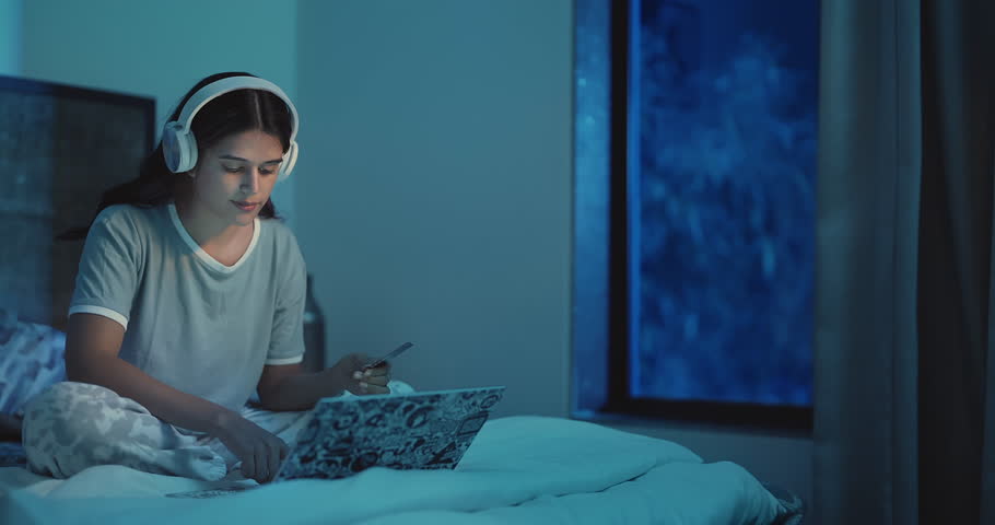 Happy Indian female sitting on bed holding credit card making online shopping payment using laptop at home. Beautiful excited woman wearing headphone purchases secure order on computer at indoor house Royalty-Free Stock Footage #1109169929