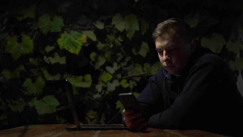 A young man at a table in a gazebo is texting on a smartphone late at night. A man in a black hoodie with a phone in his hands against a background of green grapes.