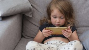 A little beautiful girl sits on the sofa in pajamas and watches cartoons on her smartphone. Child with a phone. The problem of addiction to gadgets.