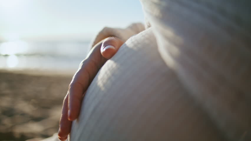 Woman hand caressing belly expecting baby close up. Unknown future mother stroking pregnant tummy sitting on sand beach at sunset. Unknown expectant mom enjoying pregnancy relaxing on evening seashore Royalty-Free Stock Footage #1109187575