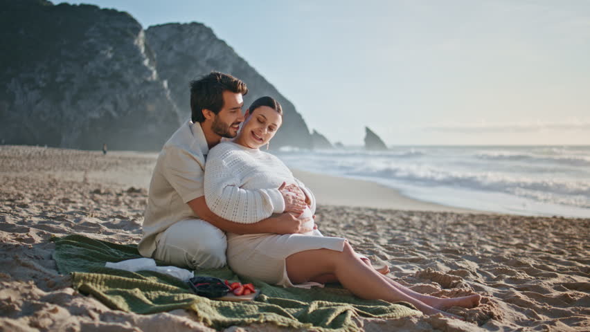 Pregnant couple sitting beach enjoying summer picnic together. Loving husband hugging big wife tummy relaxing on sunny seashore at family weekend. Calm pair sweethearts talking dreaming on seashore. Royalty-Free Stock Footage #1109187603