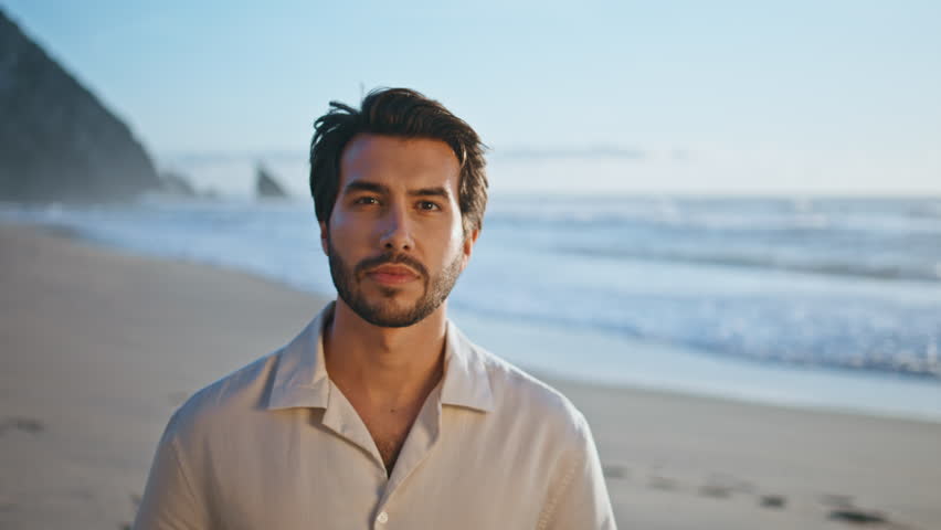 Attractive italian guy standing on beach alone enjoying beautiful seascape closeup. Portrait of handsome bearded man looking camera confidently posing on sand seashore. Hot young macho relax on coast. Royalty-Free Stock Footage #1109187611