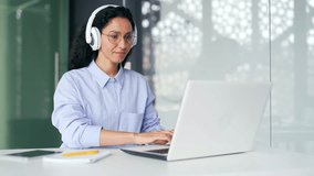 Young confident female employee in headphones typing on laptop while sitting at workplace at desk in modern office. A serious curly brunette woman works on the computer while listening to a podcast - Powered by Shutterstock - Get 15% off with code: PIKWIZARD15