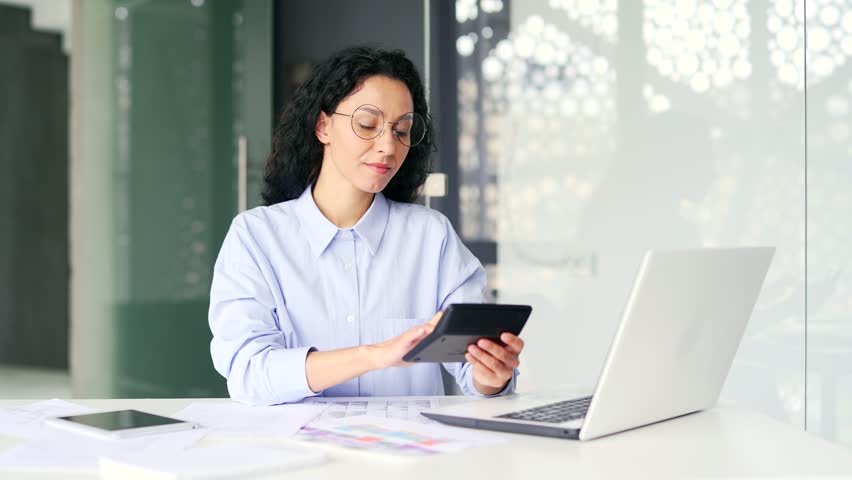Busy young female accountant forms accounts using laptop and calculator, takes notes while sitting at desk at workplace. Confident bookkeeper or financier in shirt and glasses working in modern office Royalty-Free Stock Footage #1109189045