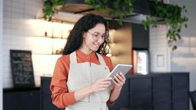 Young female small business owner uses working on a tablet while standing in a local cafe. Positive curly brunette woman employee of a pastry shop in a bathrobe reviews, checks the menu of cafeteria - Powered by Shutterstock - Get 15% off with code: PIKWIZARD15