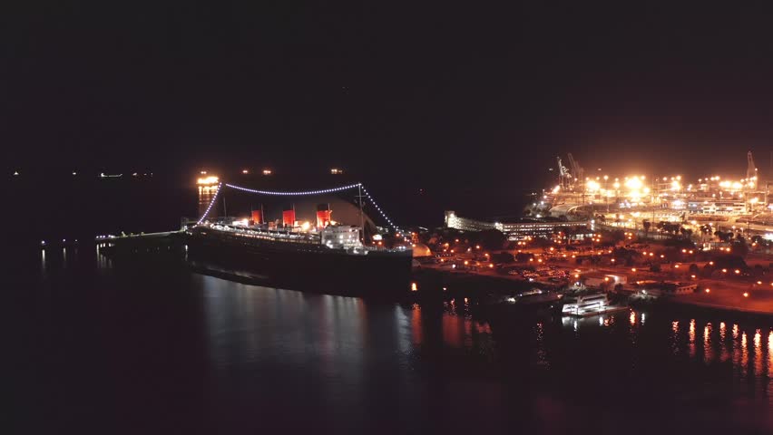 Footage of the Queen Mary II docked in Long Beach. The ship is massive and lit up festively. The water is dark and deep. The sky is dull black and the city is lit up in the background. It is night.