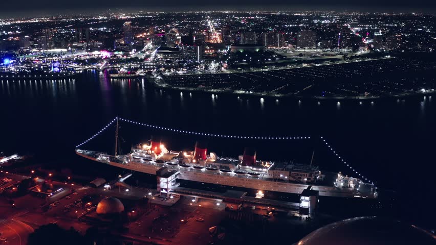 Footage of the Queen Mary II docked in Long Beach. The ship is massive and lit up festively. The water is dark and deep. The sky is dull black and the city is lit up in the background. It is night.