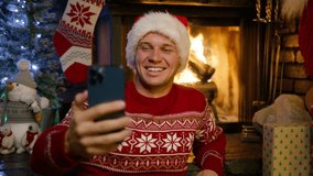 Handsome smiling man in red Santa hat winter sweater using smart phone talking to friends congratulates Merry Christmas by video call at fireplace 4K. Guy recording vlog for social media New Year 2024