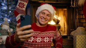 Handsome smiling man in red Santa hat winter sweater using smart phone talking to friends congratulates Merry Christmas by video call at fireplace 4K. Guy recording vlog for social media New Year 2024 - Powered by Shutterstock - Get 15% off with code: PIKWIZARD15