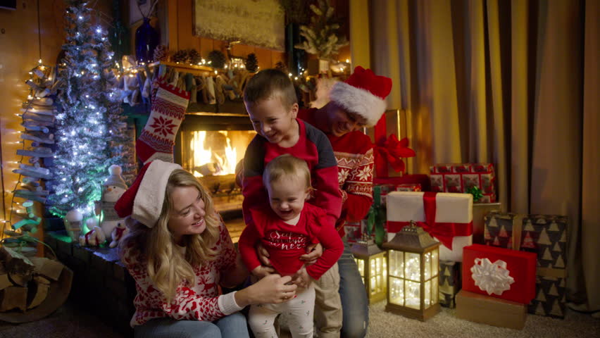 Happy family with little baby daughter, preschooler son and young beautiful parents in red Santa hats gathered together on Christmas eve near xmas tree enjoying time together at cozy fireplace RED 4K Royalty-Free Stock Footage #1109195765