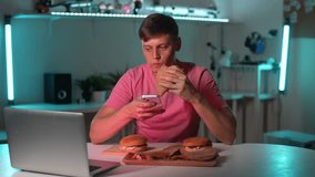 Portrait of hungry young man eating taco burger and texting on mobile phone, sitting at table with laptop and burgers during watching online movie on evening at home. Shooting in slow motion.