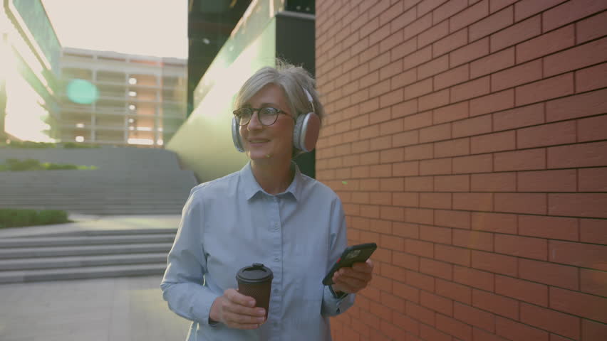 Carefree smiling senior old mature businesswoman Caucasian woman employer business female in headphones listen music browsing mobile phone walking casual going in city drink morning coffee outdoors | Shutterstock HD Video #1109197779