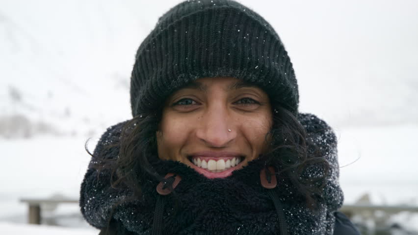 Medium close-up of Indian pretty woman with dark skin, hazel eyes, white straight teeth in black winter clothing smiling broadly at camera. Snow storm. High quality 4k footage Royalty-Free Stock Footage #1109199599