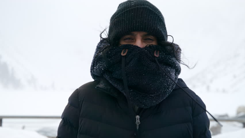 Indian woman in black winter clothing, covering mouth and nose with fur collar, squinting eyes while strong wind blows, standing outdoors during blizzard. High quality 4k footage Royalty-Free Stock Footage #1109199603