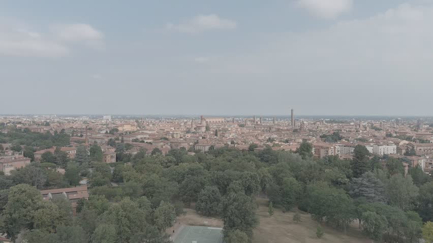 Bologna, Italy. The historic city center unfolds in a sweeping aerial panorama, revealing a dense expanse of terracotta rooftops punctuated by surviving medieval stone towers that once numbered over one hundred across the skyline. The camera glides forward over a foreground of lush green parkland before revealing the tightly packed urban fabric of the old town, where the Asinelli and Garisenda towers rise prominently among church spires and historic palazzi. The warm summer light casts soft shadows across the red-tiled roofs under a pale blue sky with scattered clouds, capturing the layered architectural heritage of one of Italy